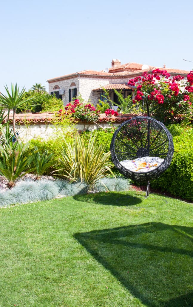 Jardin méditerranéen ensoleillé avec pelouse verte, balancelle noire et maison en pierre ornée de roses rouges sous un ciel clair.